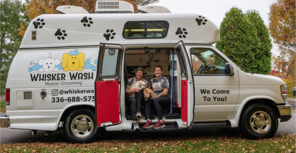Whisker Wash van with two groomers sitting in open side door holding two dogs.