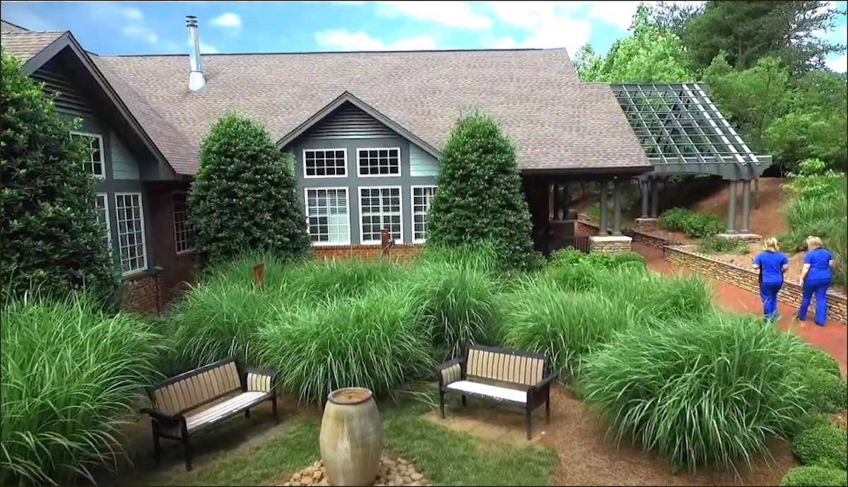 An outside view of the Kate B Reynolds Hospice Home with its large windows, one of its gardens, benches, and two staff members walking