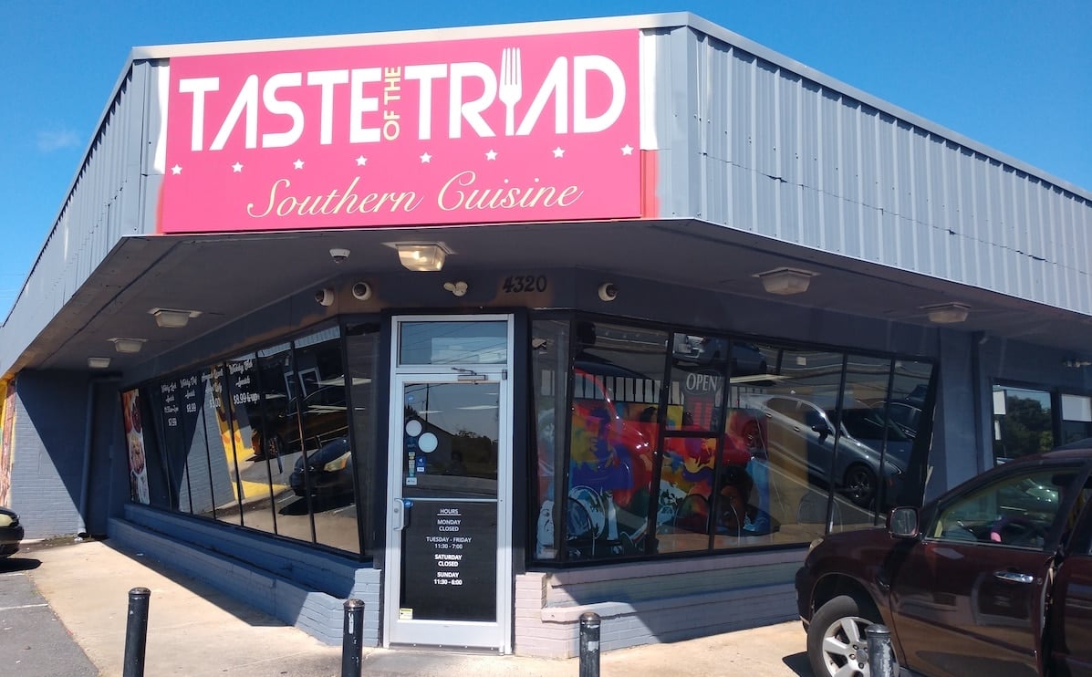 The sign says Taste of the Triad outside above the door. The i is a fork. Underneath, it says Southern Cuisine. Beneath the sign, which is on a corner on an awning, is a single glass door. There are full-length windows on either side of it. There are lights up above on the overhang.