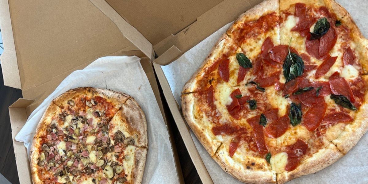 two pizzas in boxes covered with toppings 